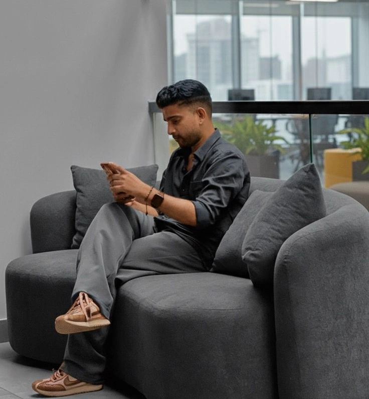 A man sitting on a modern grey sofa in a contemporary office space looking at his phone while dressed in a dark shirt and grey pants best freelance digital marketing expert in malappuram